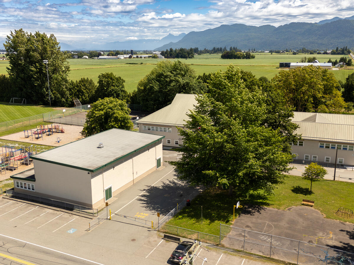 Exterior image of an elementary school that sits in the farm land of Abbotsford. School field, playground cand be seen, along with cloudy sky and mountain range in the distant background.